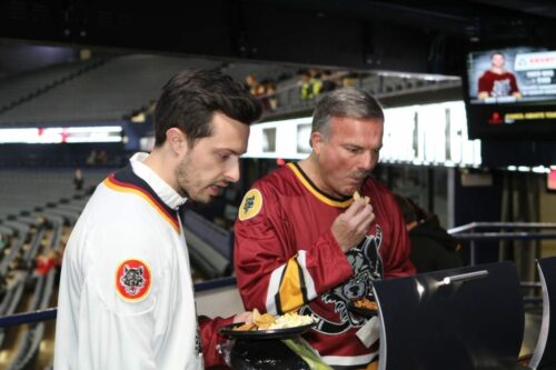 Two men in Chicago Wolves hockey jerseys stand at a concession stand inside a sports arena during the 2018 March Networking Night. One man is eating nachos from a plate, while the other watches. The arena’s seating area is visible in the background, along with some illuminated screens.