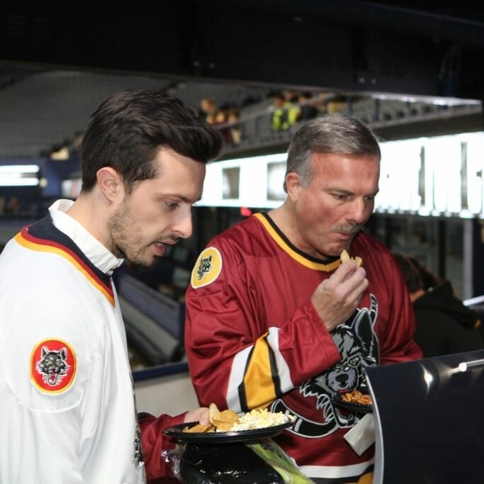 Two men in Chicago Wolves hockey jerseys stand at a concession stand inside a sports arena during the 2018 March Networking Night. One man is eating nachos from a plate, while the other watches. The arena’s seating area is visible in the background, along with some illuminated screens.