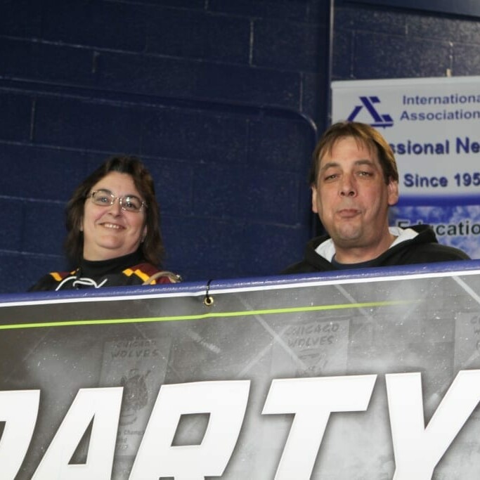 Two people are standing behind a large banner with the word "PARTY" on it. They are in an indoor venue with dark blue walls. Above and to the right of the individuals, there's a sign for the International Air Cargo Association of Chicago's 2018 March Networking Night, supported by Chicago Wolves.