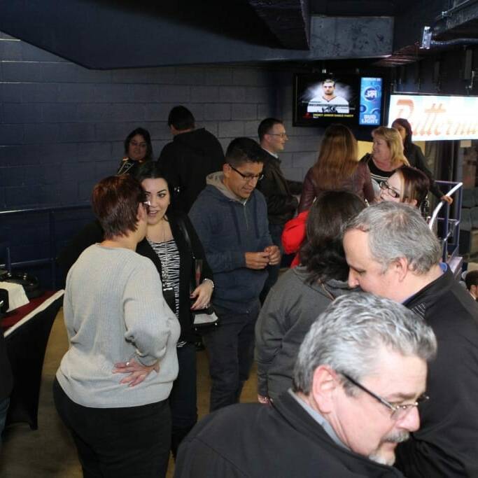 People are gathered indoors at Networking Night, socializing and gesturing while others are seated. The partially visible buffet table to the left tempts attendees. In the background, a monitor displays a man talking, with a brightly lit sign partially visible above the seating area from 2018 March's Chicago Wolves event.