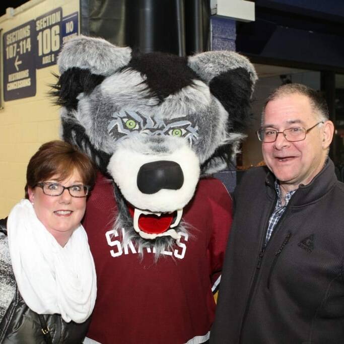 A woman in a white scarf and glasses and a man in a grey jacket pose with someone in a large grey and black wolf mascot costume. The group is standing indoors near a sign for section 106 and other arena signage at the Chicago Wolves' March Networking Night, 2018.