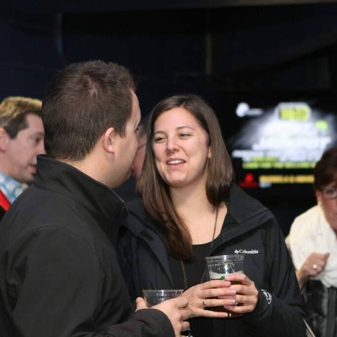 A man and woman wearing black jackets are engaged in a conversation, each holding a drink, at the 2018 Chicago Wolves Networking Night. Behind them, another man in a black jacket looks on, while a woman in a white outfit drinks from a cup. A screen displaying advertisements is visible in the background.