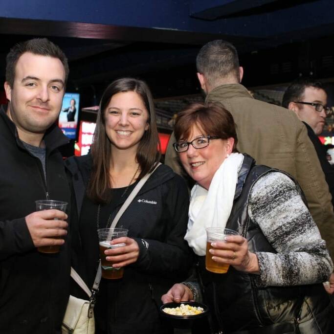 Three people stand together indoors at the 2018 March Networking Night, smiling and holding drinks. The man on the left is wearing a dark jacket, the woman in the middle is in a black jacket, and the woman on the right is wearing glasses, a white scarf, and a patterned jacket. People and screens are visible in the background.