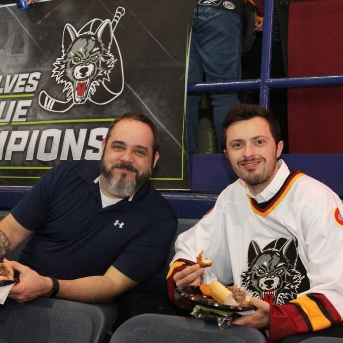 Two men are sitting in an arena enjoying food. The man on the left wears a navy polo shirt, while the man on the right sports a Chicago Wolves hockey jersey. Behind them is a banner that reads, "Chicago Wolves V League Champions," featuring the team logo from Networking Night, 2018 March.