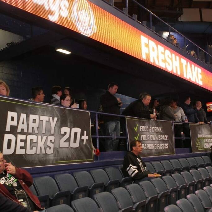 A group of people are standing and sitting in the stands of an indoor arena during the 2018 March Networking Night hosted by the Chicago Wolves. A banner with "Party Decks 20+" is displayed prominently, along with other banners advertising food, drink, and parking passes. Some spectators are focused on the events happening in the arena.