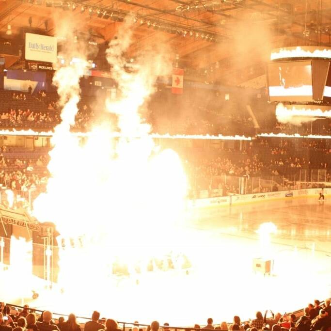 A large indoor ice hockey arena is pictured with many spectators. There is a bright explosion or pyrotechnic display occurring near one end of the rink, casting a yellow glow over the ice and crowd. This mesmerizing spectacle seems to be part of a Night Chicago Wolves event.