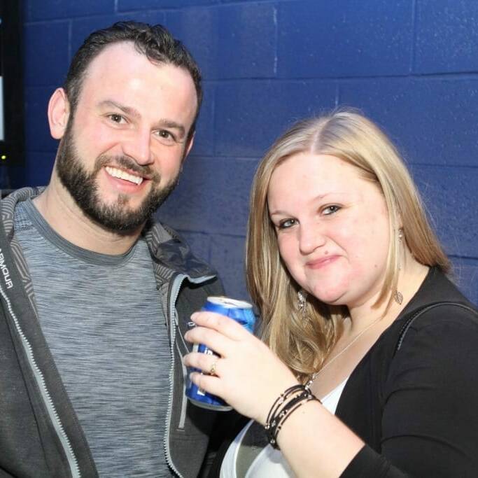 Two people posing together against a blue brick wall. The person on the left, sporting short dark hair and a beard, is wearing a gray shirt and dark jacket. The person on the right has long blonde hair, holding a can of drink, and is dressed in black and white attire at the 2018 March Networking Night for Chicago Wolves fans.