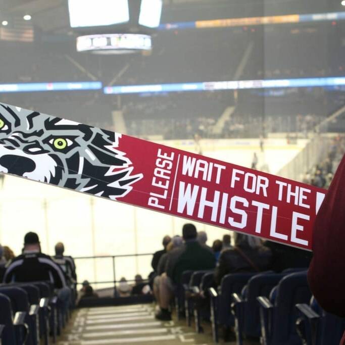 A person holding a sign with a fierce wolf face that reads "PLEASE WAIT FOR THE WHISTLE" at an indoor hockey arena during the 2018 March Networking Night hosted by the Chicago Wolves. People are seated and watching a game on the ice rink in the background.