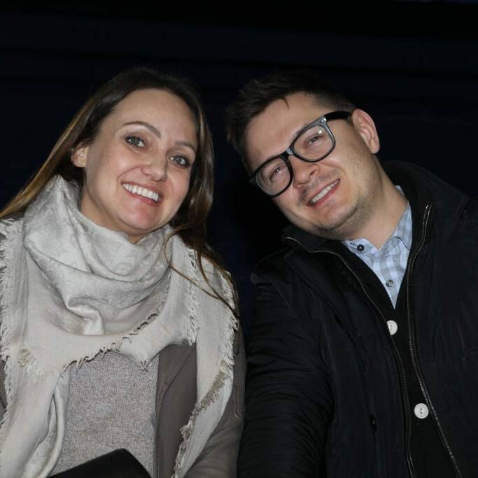 Two people are smiling and looking at the camera. The person on the left is wearing a light-colored scarf and a jacket, while the person on the right is wearing glasses, a dark jacket, and a collared shirt. The background is dark. It was captured during the 2018 March Networking Night with the Chicago Wolves.