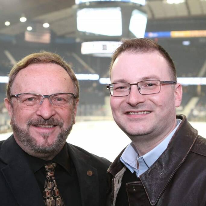 Two men are smiling at the camera in this indoor arena during the 2018 March Networking Night. The man on the left is wearing glasses, a dark suit, and a patterned tie, while the man on the right is sporting glasses, a brown leather jacket, and a blue collared shirt. The arena is brightly lit in the background.