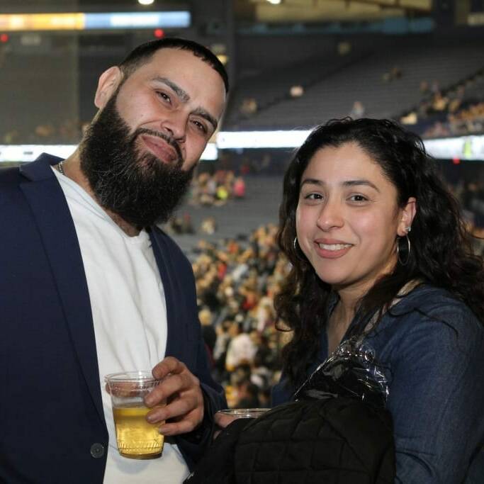 A man with a beard and a woman with long curly hair are smiling while standing together in a large indoor arena during Networking Night. The man is holding a drink in a plastic cup. The background shows a blurred crowd seated in rows of bleachers, evoking memories of the 2018 March Chicago Wolves event.