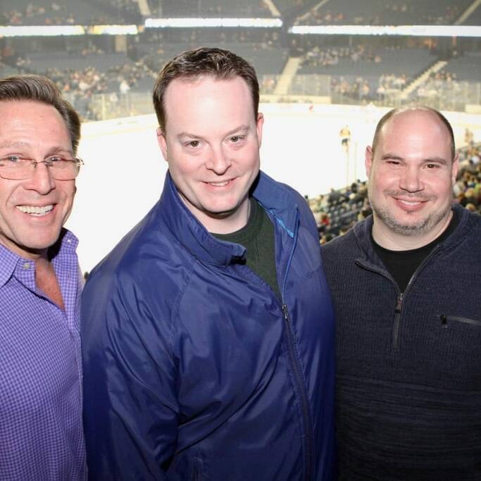 Three men are smiling and posing for a photo at an indoor stadium during the Chicago Wolves' 2018 March Networking Night. The man on the left is wearing a purple shirt, the man in the middle is wearing a blue jacket, and the man on the right is in a dark grey sweater. The stadium seats and ice rink are visible in the background.