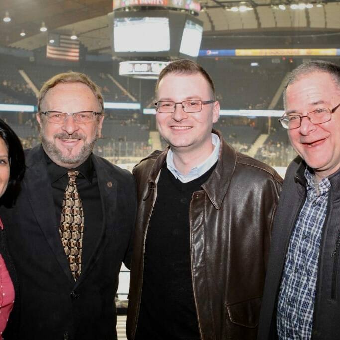 Four people are standing together and smiling at Networking Night 2018. The group includes two men in the middle, one in a leather jacket and the other in a suit, with a woman on the far left and another man in a casual fleece jacket on the far right. The background shows stadium seating at a Chicago Wolves game.