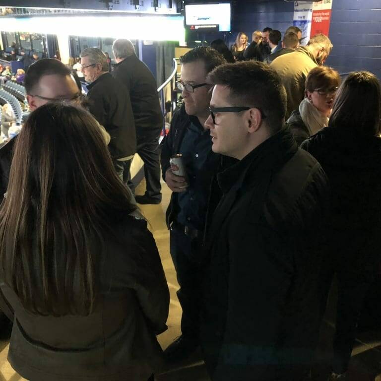 A group of people stands in an indoor area with stadium seating and spectators visible in the background. They appear to be engaged in conversation during a Networking Night at a Chicago Wolves game. The lighting is dim, and some individuals hold drinks, suggesting a social event or gathering at a sports venue.