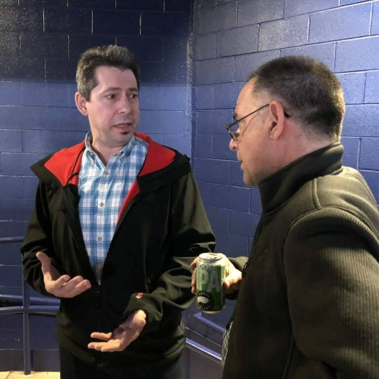 Two men engage in conversation indoors against a blue brick wall. The man on the left wears a black jacket with red lining and a blue checkered shirt, gesturing with his hands. The man on the right, holding a can of IPA beer, wears glasses and a dark jacket—it's clear they're enjoying Networking Night after a Chicago Wolves game.