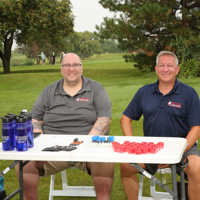 Two men are seated at a table outdoors on a golf course during the 2021 IACAC Golf Outing. The table holds blue water bottles, red containers, utensils, and plastic cups. Both men are smiling and wearing shirts with logos. Trees and greenery are visible in the background.