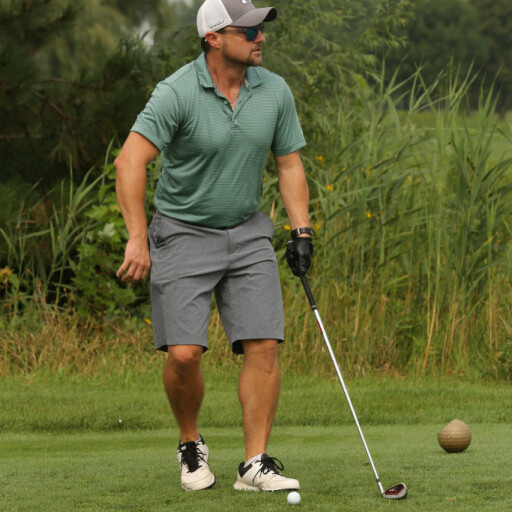 A man wearing a green polo shirt, gray shorts, a gray cap, and dark sunglasses is preparing to take a golf swing on a lush green course during the IACAC Golf Outing 2021. He holds a golf club in one hand and stands beside a golf tee and ball, surrounded by tall grass and trees in the background.