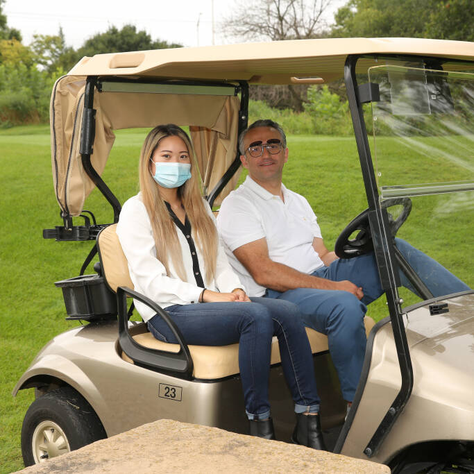 A woman wearing a medical mask and a man with sunglasses are seated in a beige golf cart on a lush green golf course during the IACAC Golf Outing. The woman wears a white shirt and blue jeans, and the man wears a white polo shirt and blue pants. Trees and greenery are visible in the background.