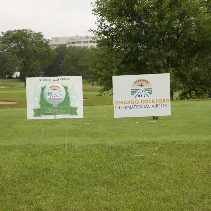 Two signs on a grassy field. The left sign reads "GOLF OUTING 2023 SPONSOR" and features a logo. The right sign reads "CHICAGO ROCKFORD INTERNATIONAL AIRPORT" with a logo. Trees and a building are visible in the background, reminiscent of the successful IACAC Event held here.