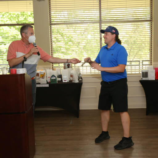 Two men are in a room with large windows and blinds. One man, in a mask and pink shirt, stands behind a podium, holding a microphone and a small item. The other man, in a blue shirt and cap, is smiling and extending his hand to receive the item—a scene from the IACAC 2021 Golf Outing.