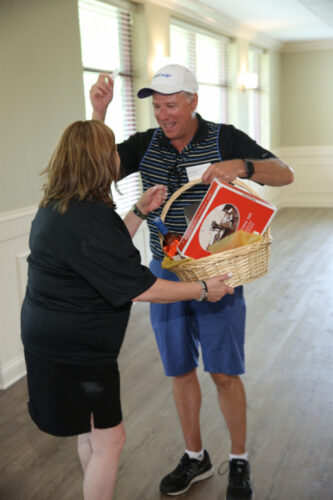 A man in casual sports attire, holding a gift basket and an envelope, smiles and interacts with a woman dressed in black. They are in a room with large windows, wood flooring, and soft green walls. The woman’s back is to the camera as they share a joyful moment reminiscent of IACAC's 2021 Golf Outing.