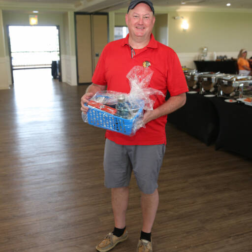 A man wearing a red polo shirt and a black cap is holding a blue basket filled with wrapped items. He is standing on a wooden floor in a room with tables set up in the background, likely for the 2021 IACAC Golf Outing event or gathering.