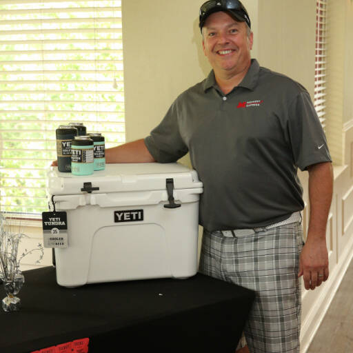 A man in a polo shirt, baseball cap, and plaid shorts stands indoors beside a table displaying a YETI cooler and YETI-branded drinks. Smiling with one arm resting on the cooler, he appears ready for the 2021 IACAC Golf Outing. A window with blinds is in the background.