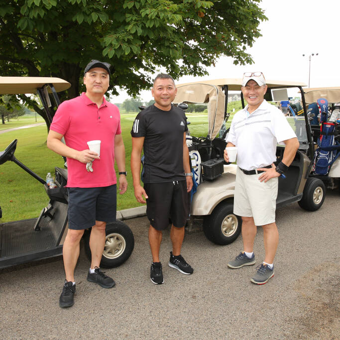 Three men stand in front of a row of golf carts at the 2021 IACAC Golf Outing. The man on the left wears a pink polo shirt and black shorts, the middle man sports a black athletic outfit, and the man on the right dons a white polo with light-colored shorts. They smile and hold drinks.