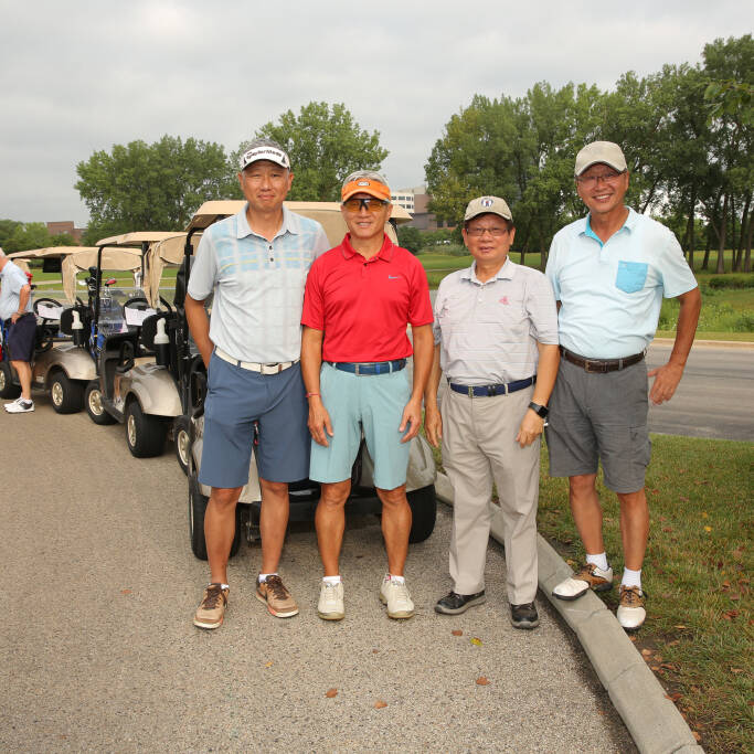 Four men are standing in a row on a golf course, all casually dressed in golf attire during the 2021 IACAC Golf Outing. Behind them are golf carts lined up on a paved path. The background features lush green trees and a cloudy sky. The mood appears to be relaxed and friendly.