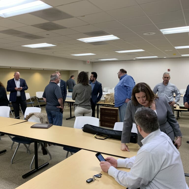 In April 2019, a group of people are engaged in conversations and using devices in a meeting room. The room has tables, chairs, and a clock on the wall. Some individuals are standing around a table with food and beverages from the luncheon, while others are seated or walking.