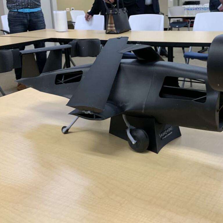 A small, sleek black model airplane with a unique design is placed on a wooden table at the April 2019 luncheon. The aircraft has vertical wings and jet engine thrusters at the rear. Several people and chairs are visible in the background in a bright room.