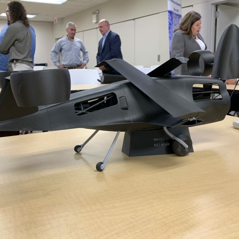 A detailed scale model of a futuristic aircraft sits on a conference room table during the April luncheon in 2019. The aircraft features a sleek black design with vertical and horizontal stabilizers and ducted fan engines. In the background, several people are engaged in conversation.