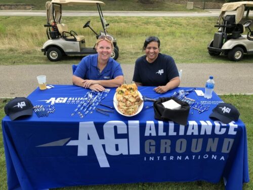 Two people sit behind a table covered with promotional items and sandwiches at the 2018 IACAC Golf Outing. The tablecloth features the logo and name "AGI Alliance Ground International." Golf carts are visible in the background.