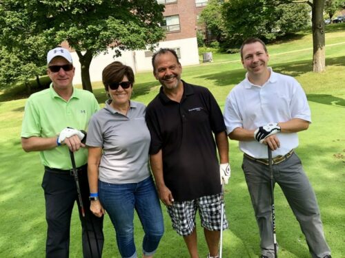 Four people stand together on a golf course during the 2018 IACAC Golf Outing, smiling at the camera. One person is on the left wearing a light green shirt and sunglasses. The others are dressed in casual golf attire, with clubs in hand. Trees and a building are visible in the background.