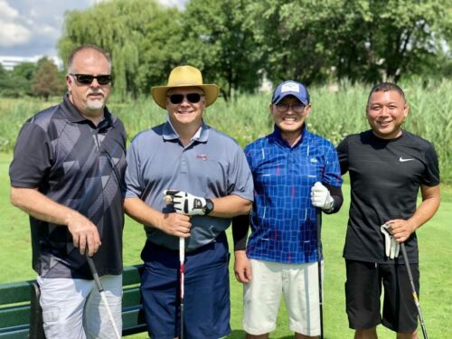 Four men are standing together on a golf course at the 2018 IACAC Golf Outing, all holding golf clubs. They are smiling and dressed in different golf attire, including polo shirts, shorts, and hats. Trees and grass are visible in the background under a sunny sky.