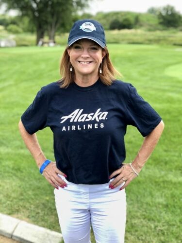 A woman stands on the grass, smiling during an IACAC outing. She wears a navy blue "Alaska Airlines" t-shirt, white pants, and a navy baseball cap. Her hands are on her hips as trees and a cloudy sky form the backdrop.