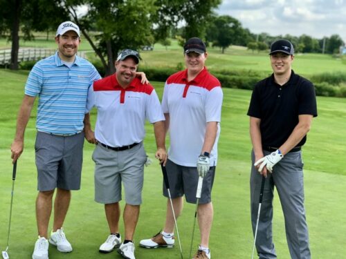 Four men are standing together on a golf course during the IACAC 2018 Golf Outing, each holding a golf club. They are smiling and dressed in golf attire, including polo shirts, shorts, and caps. The background features lush green grass, trees, and a bright sky with some clouds.