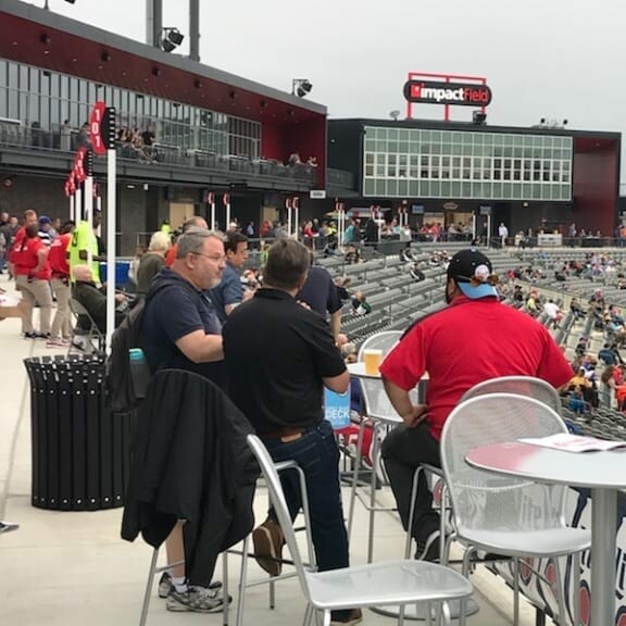 A group of people sitting at a table with barstools in an outdoor stadium seating area, with rows of seats and spectators in the background. The venue is bustling with activity from the 2018 IACAC Baseball Networking Event, and there is a large sign that reads "impact field" visible in the background.