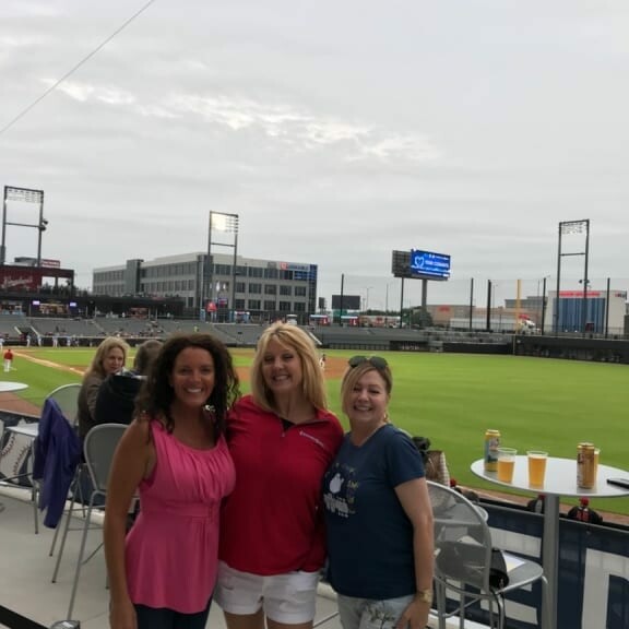 Three women stand together smiling in front of a baseball field with green grass and stadium seating in the background. They are casually dressed, and a few drinks are on the table behind them. It's an overcast day at the 2018 IACAC Baseball Networking Event, and the atmosphere appears relaxed and friendly.