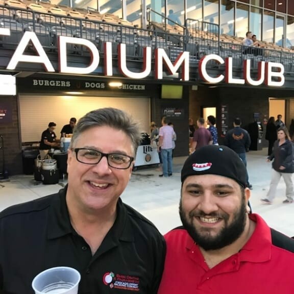 Two men smiling at the camera in front of a venue labeled "Stadium Club". The man on the left, holding a drink and wearing glasses and a black shirt, seems thrilled about the baseball event. The man on the right, in a red shirt and black cap, radiates excitement. People are visible in the background.