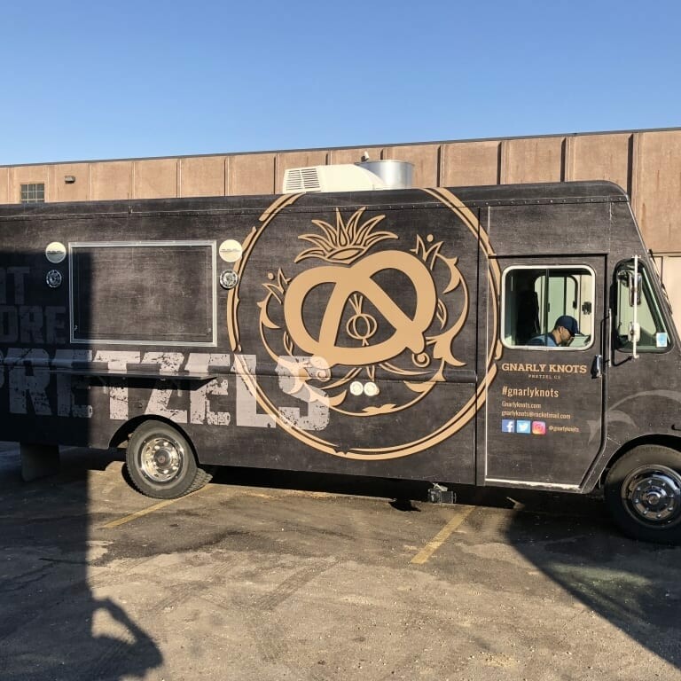 A black food truck with the Gnarly Knots logo and the tagline "Eat More Pretzels." The truck also displays the social media handles and icons for Facebook, Twitter, and Instagram (@gnarlyknots). The truck is parked in a sunlit parking lot beside a beige building, ready to serve delicious treats at September's Oktoberfest.