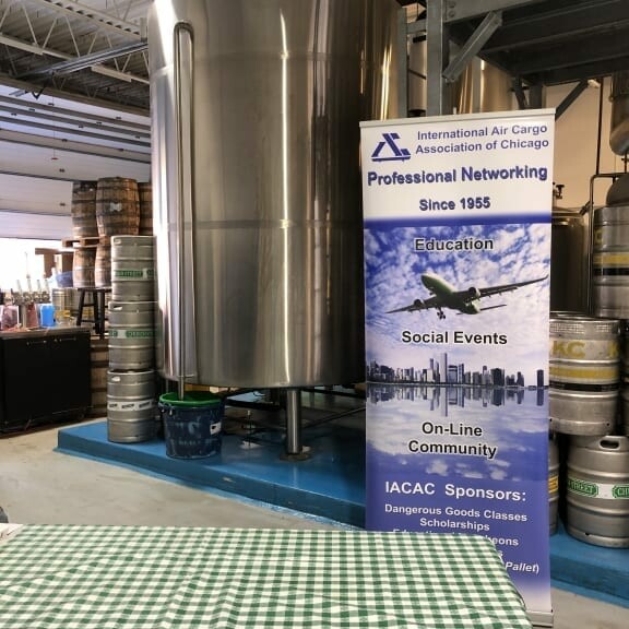 A large stainless steel tank stands in a spacious, industrial indoor area. In front of it is a banner for the International Air Cargo Association of Chicago (IACAC), promoting professional networking, education, and social events. To celebrate Oktoberfest in September, a green-and-white checkered tablecloth covers a nearby table.