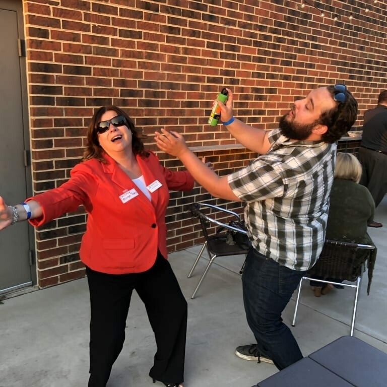 Two people are playfully interacting outside near a brick wall. One person, wearing a red jacket and sunglasses, poses exuberantly with outstretched arms, reminiscent of the lively energy at September Oktoberfest, while the other person, in a plaid shirt, leans back, pretending to be startled.