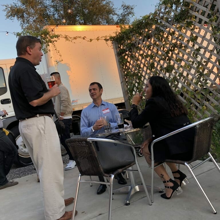 A group of people are gathered outdoors near a wooden lattice during the 2018 IACAC. Four individuals are visible; two are seated at a metal table, one is standing and talking, and another is in the background. A white delivery truck is parked nearby, while string lights hang above, adding an Oktoberfest charm.