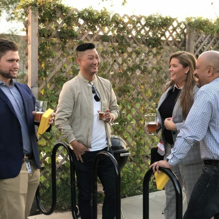 A small group of people, reminiscing about the 2018 IACAC, gather outside, conversing and laughing. One man gestures while talking, another holds a beer reminiscent of September Oktoberfest, and others listen and smile. They stand near a wooden trellis with green vines as the sun sets in the background.