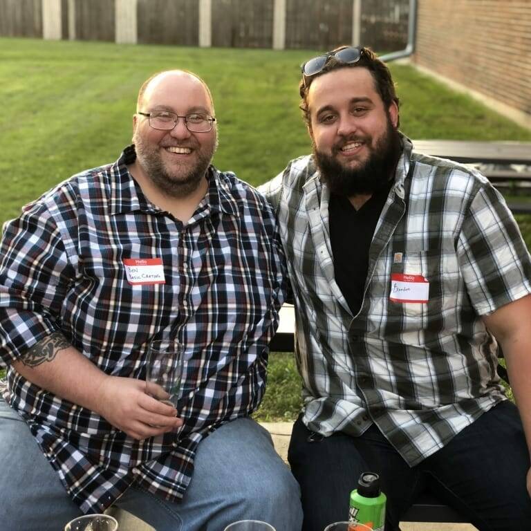 Two men are sitting together at the September Oktoberfest, wearing checkered shirts and name tags. Both are smiling at the camera, with one man holding a drink. A grassy area and wooden fence are visible in the background, capturing the essence of this memorable IACAC 2018 gathering.