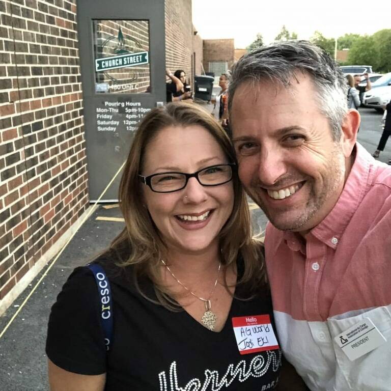 A man and woman smile for a selfie outside a building during the 2018 IACAC event. The woman, wearing glasses and a black shirt with white text, has a name tag on her chest. The man in the light blue shirt also sports his name tag. They are standing near a brick wall.