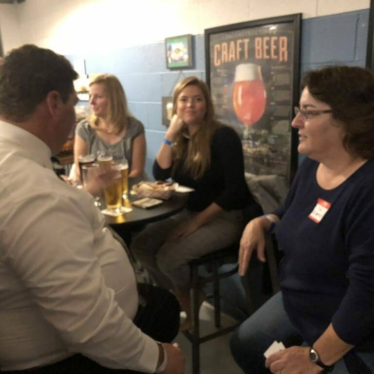 A group of people sits and stands around a table in a casual setting, reminiscent of an early September gathering. They are holding drinks and engaged in conversation. A framed "Craft Beer" poster is visible on the wall behind them, lending an Oktoberfest vibe to the relaxed and friendly atmosphere.