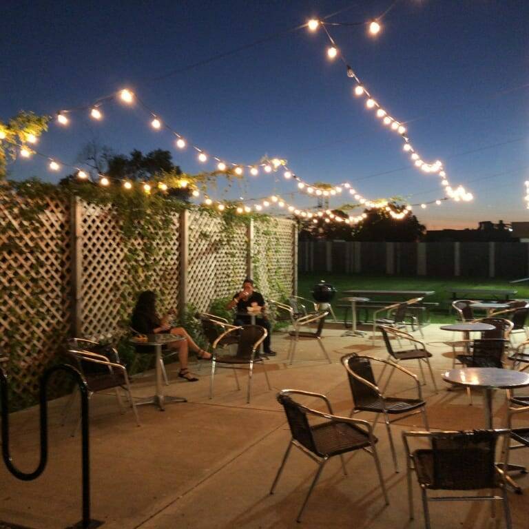 A cozy outdoor patio with string lights overhead, creating a warm atmosphere at dusk during September. Two people are seated at separate tables, engaging in conversation. The patio is bordered by a wooden lattice fence adorned with greenery, and several empty tables and chairs are visible.