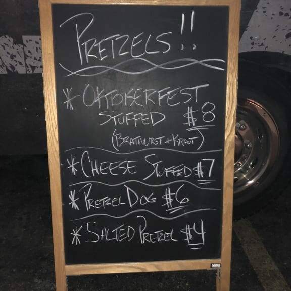 A chalkboard menu reads: "PRETZELS!!" at the top. Below, it lists: "Oktoberfest Stuffed (Bratwurst & Kraut) $8," "Cheese Stuffed $7," "Pretzel Dog $6," and "Salted Pretzel $4." The board is framed in wood and placed in front of a food truck at night,